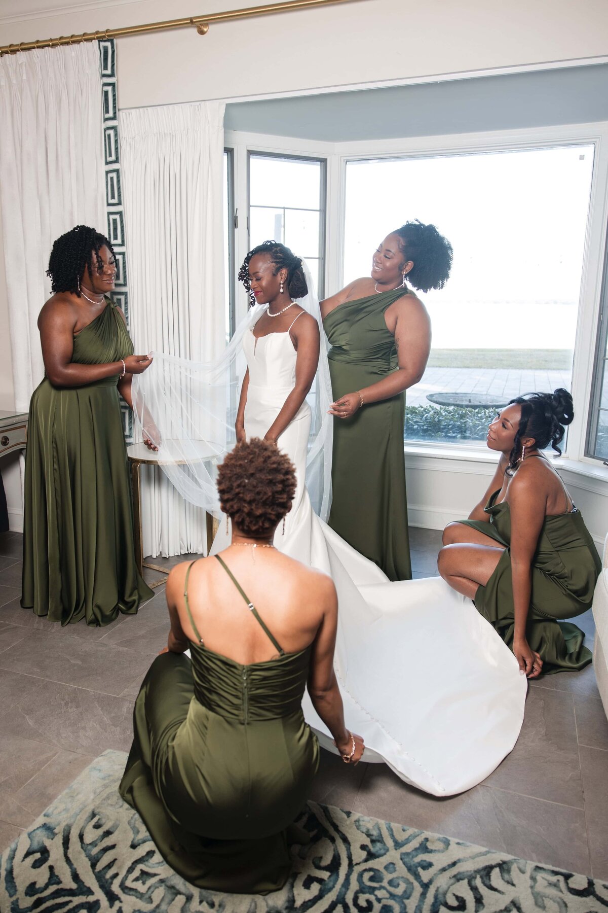 bride stands gracefully in her wedding gown as her bridesmaids, dressed in olive green, help adjust her veil and train before the ceremony at Azaleana Manor in Orange Park, Florida. Natural light streams in through large windows overlooking the river, creating a serene and elegant atmosphere. The moment is filled with love, laughter, and anticipation, beautifully captured by a Jacksonville wedding photographer known for documenting real, timeless moments with style and care.