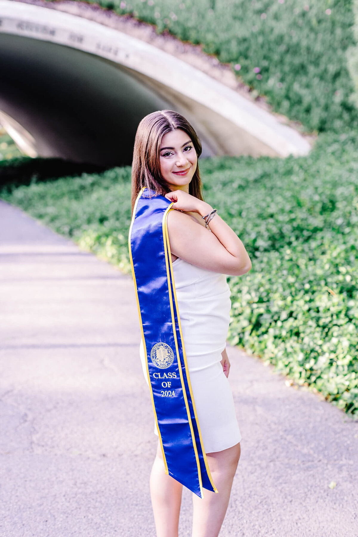 A young woman in a white dress stands on a path, wearing a blue graduation sash with Class of 2024 written on it. She smiles at the camera with greenery and an arched tunnel in the background.