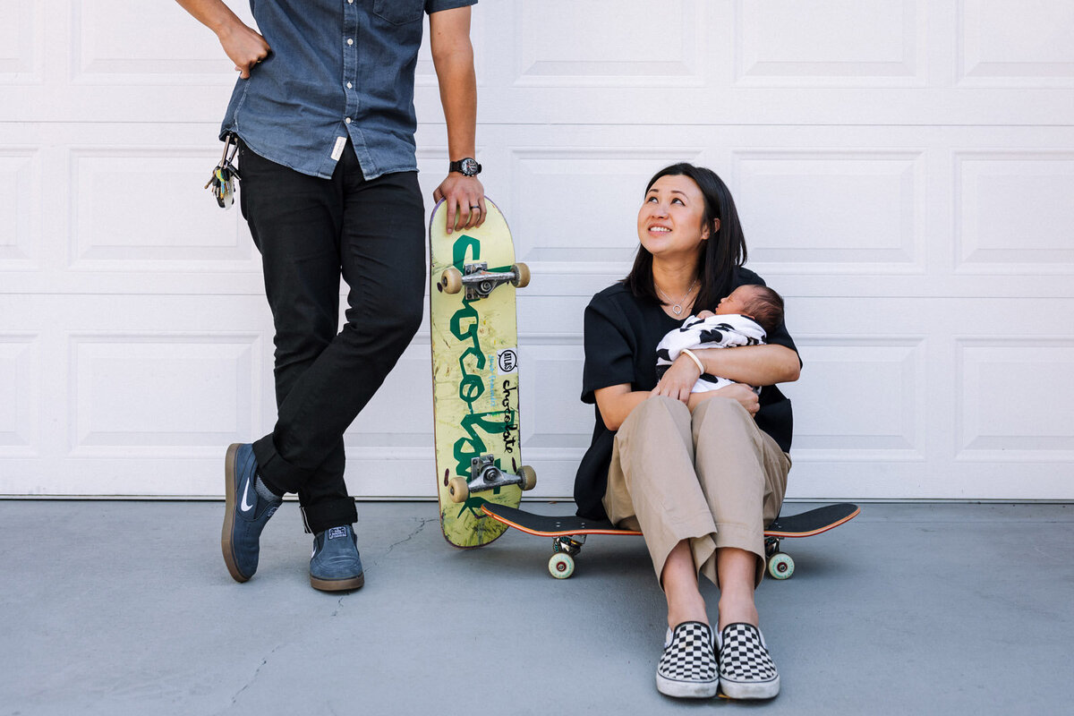 mom sitting on skatekboard wearing checkered vans holding newborn and dad stands next to her leaning on his skateboard