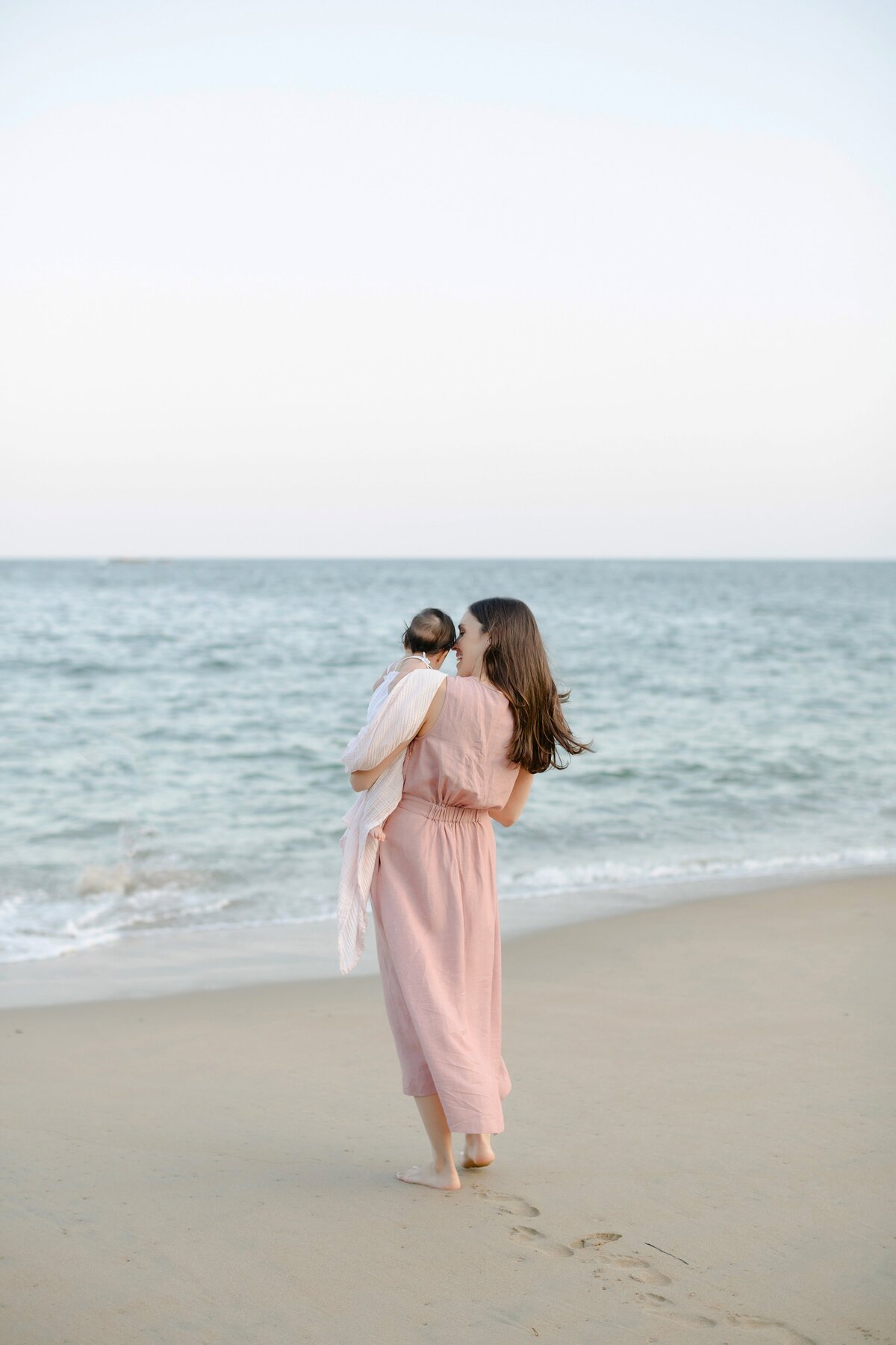 Woman holds child while walking on the beach in a pink dress.