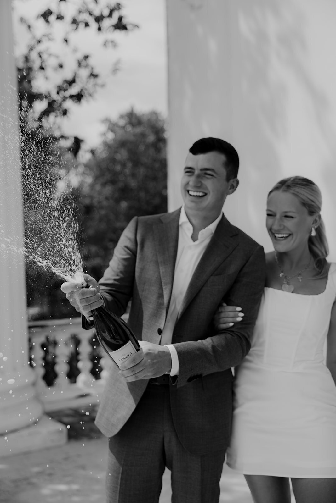 Man sprays bottle of champagne. Woman holding to his arm and laughts.
