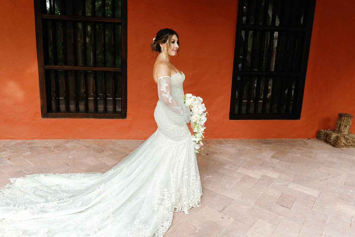 Victoria poses against a rustic red wall in Cartagena, showcasing her lace wedding gown and cascading bouquet.