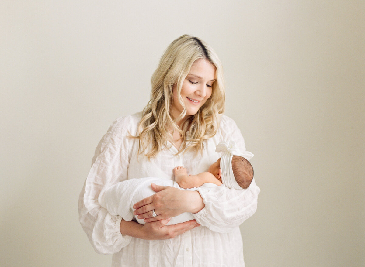 New mom stands in her room and looks lovingly down at her baby. Image by Raleigh newborn photographer A.J. Dunlap Photography.