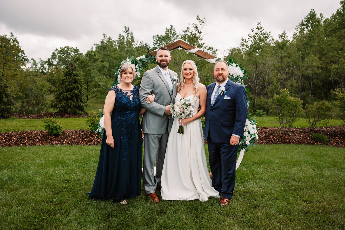 Newlyweds stand under the arbor smiling with mom and dad on either side