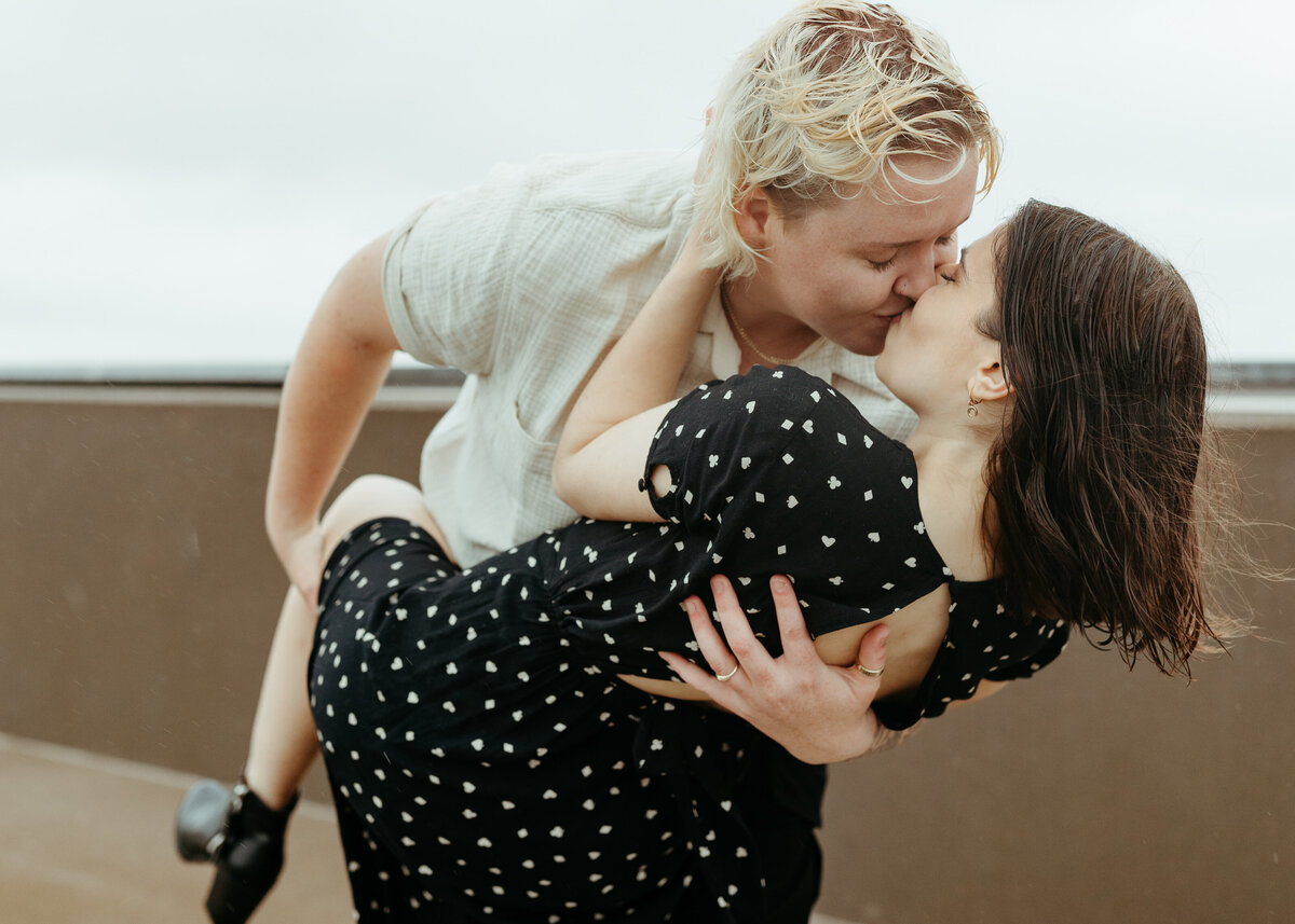 A person lifts another in a spontaneous kiss, set against a cloudy sky on an urban rooftop