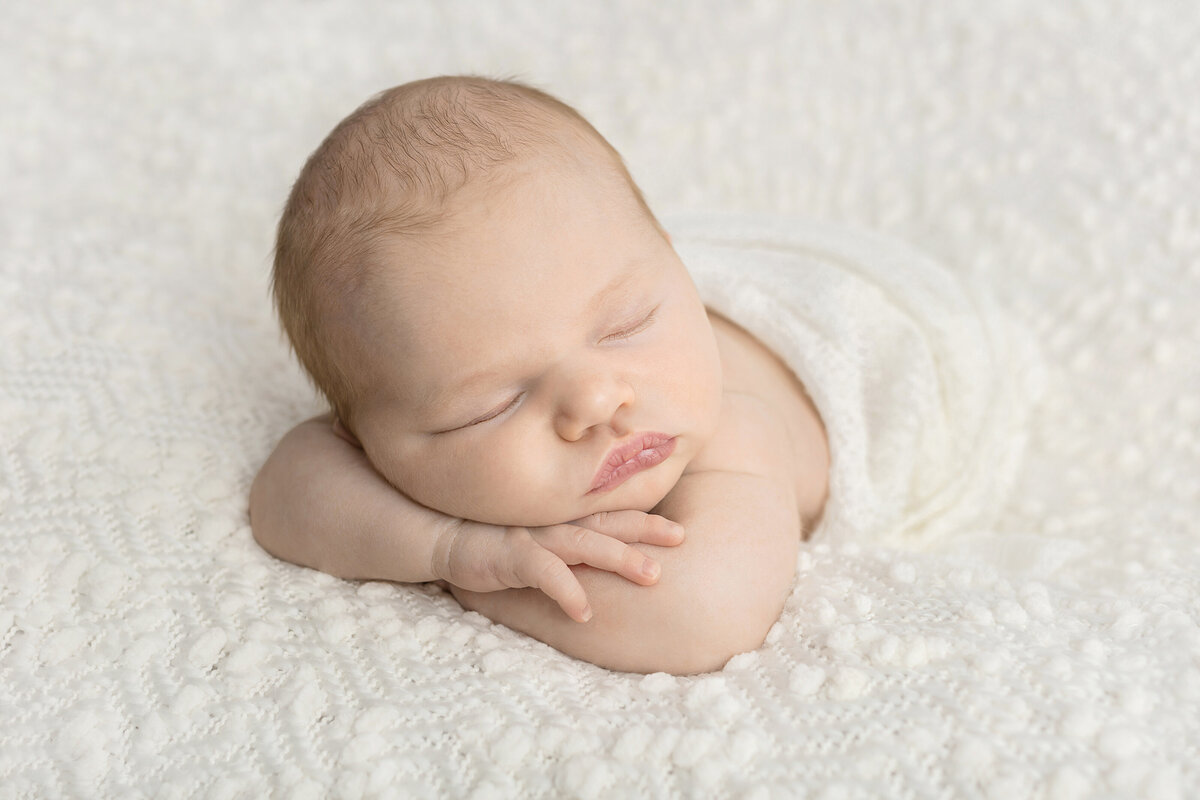 A sleeping newborn with feathery hair and slightly protruding lips is pictured sleeping on their crossed arms. They are lightly swaddled in a creamy knit blanket and are lying on a chunky, patterned ivory colored blanket.