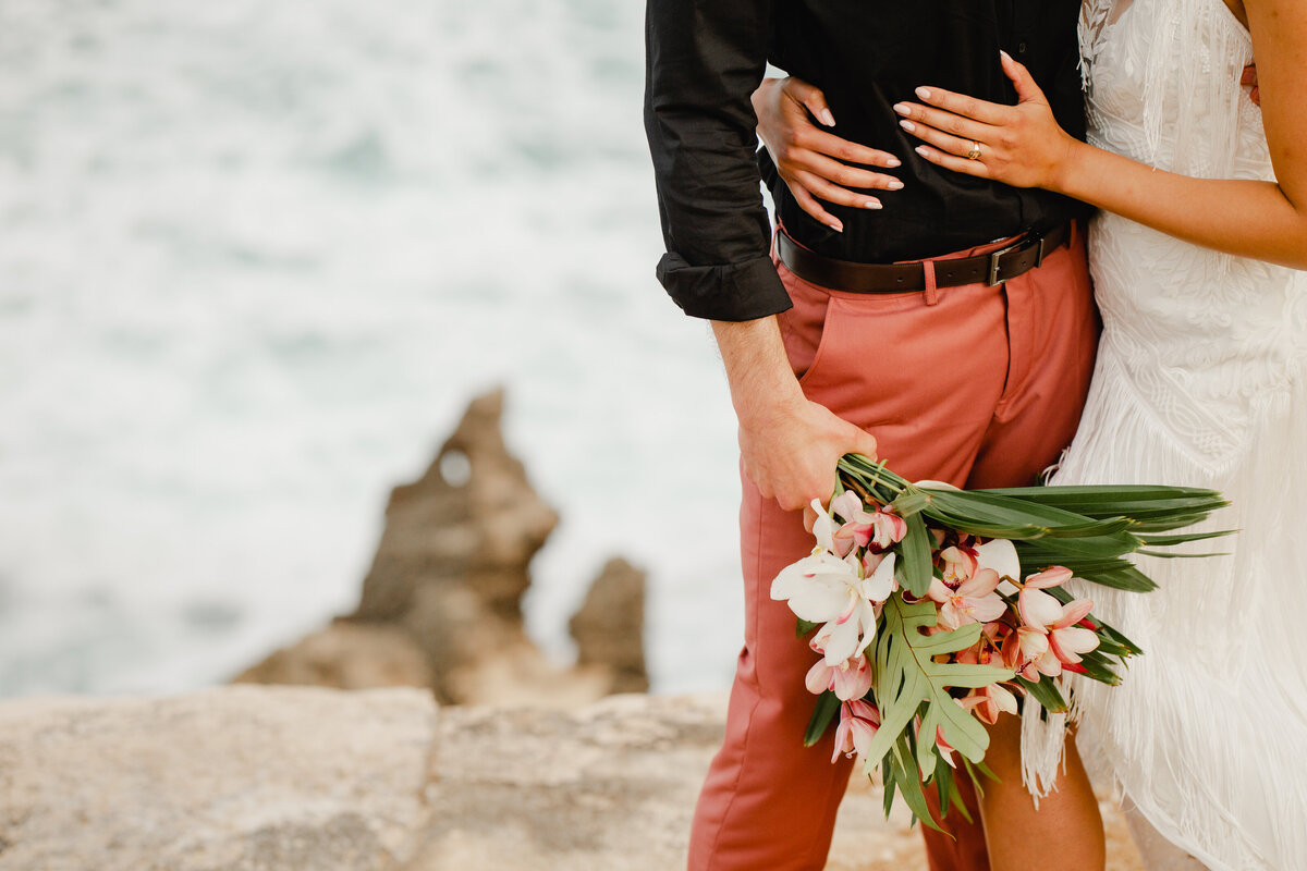 A detail photo of a bride with her arms around her groom's waist. He is holding her beautiful bouquet of flowers and they are standing on a beach in Hawaii.