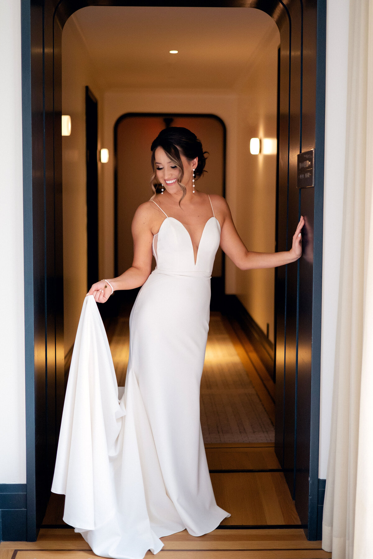 A photo of a bride holding the tail of her chiffon wedding gown and looking at her dress in her hand.  She is smiling and standing in a black rimmed doorway