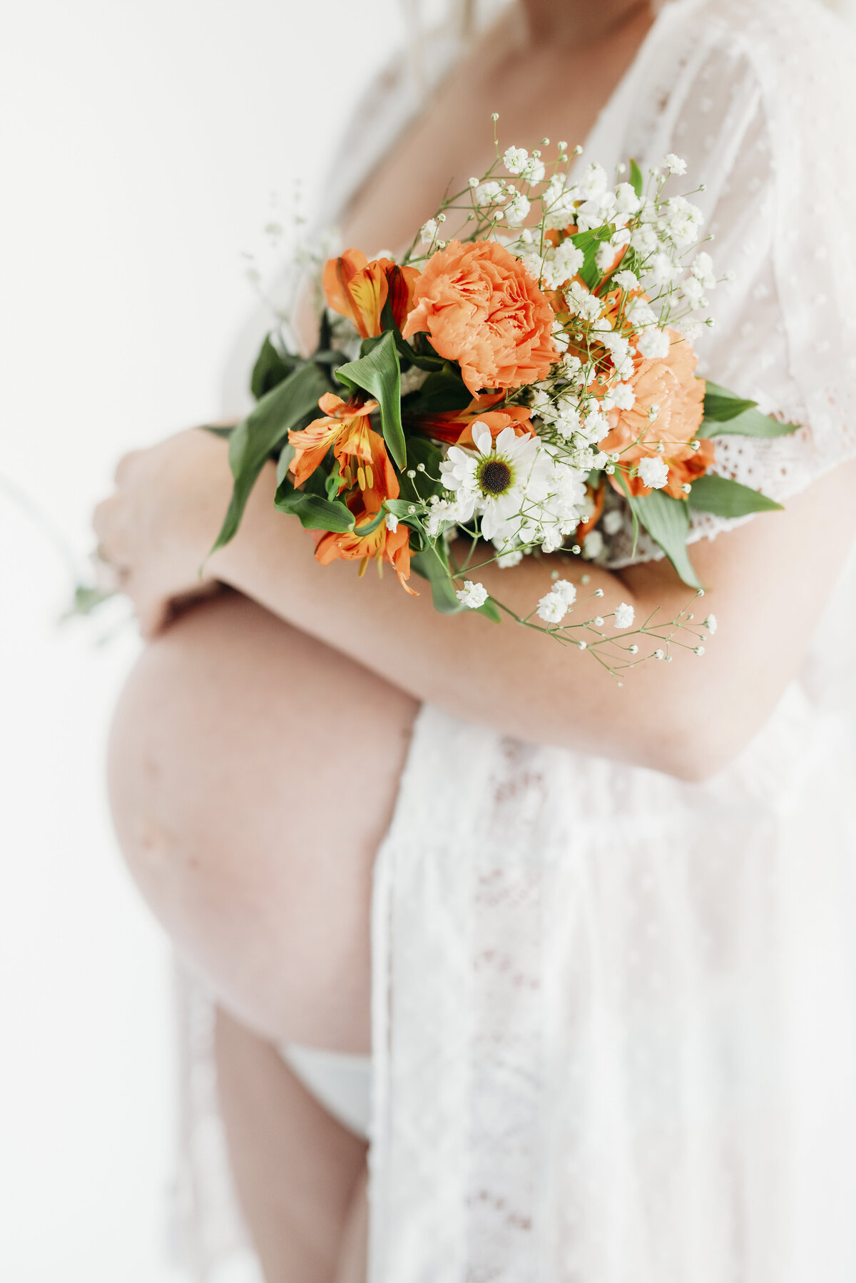 Pregnant woman in a sheer lace robe holding a bouquet of orange and white flowers over her belly.