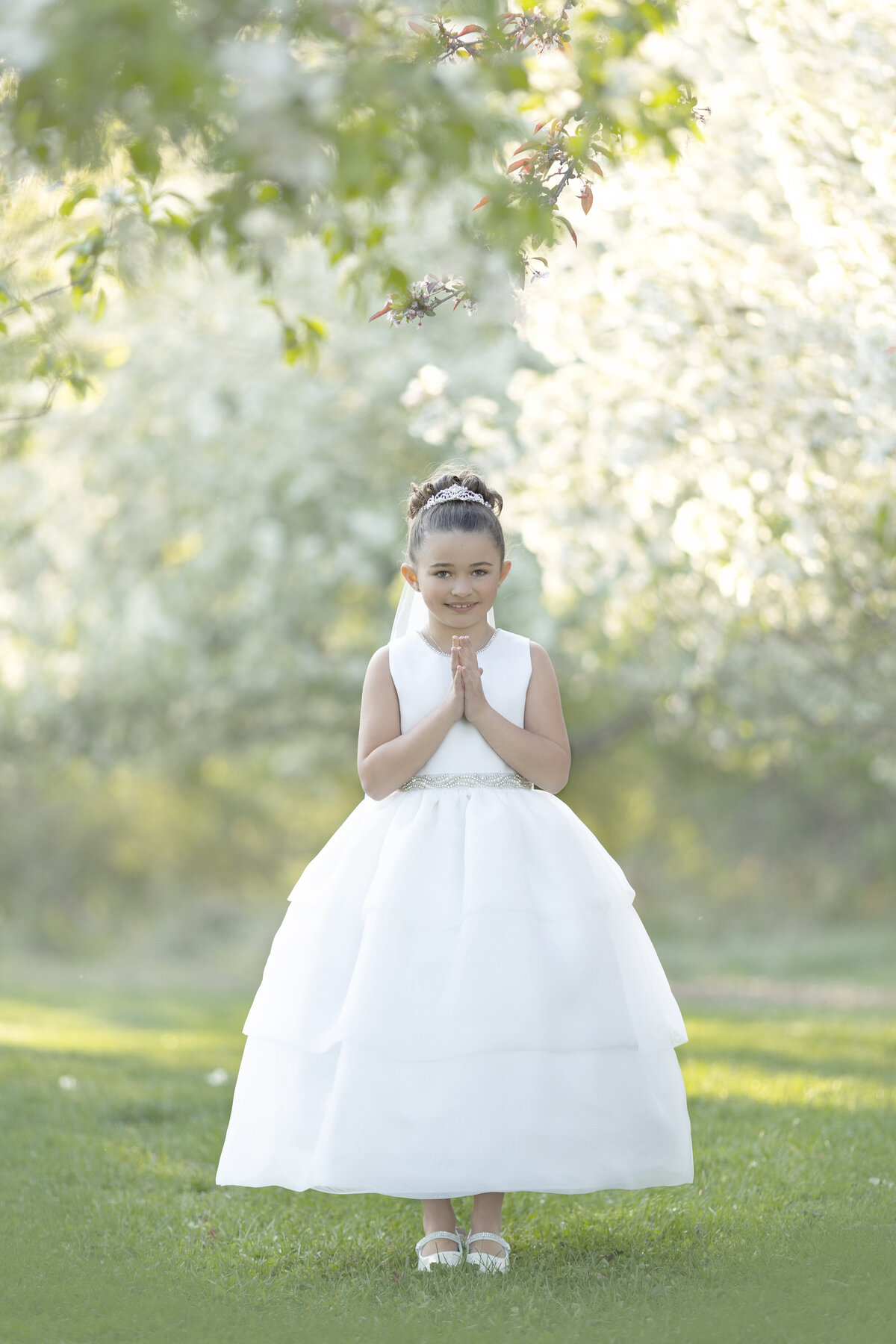 A young girl holds her hands in prayer in a white dress in a park at sunset
