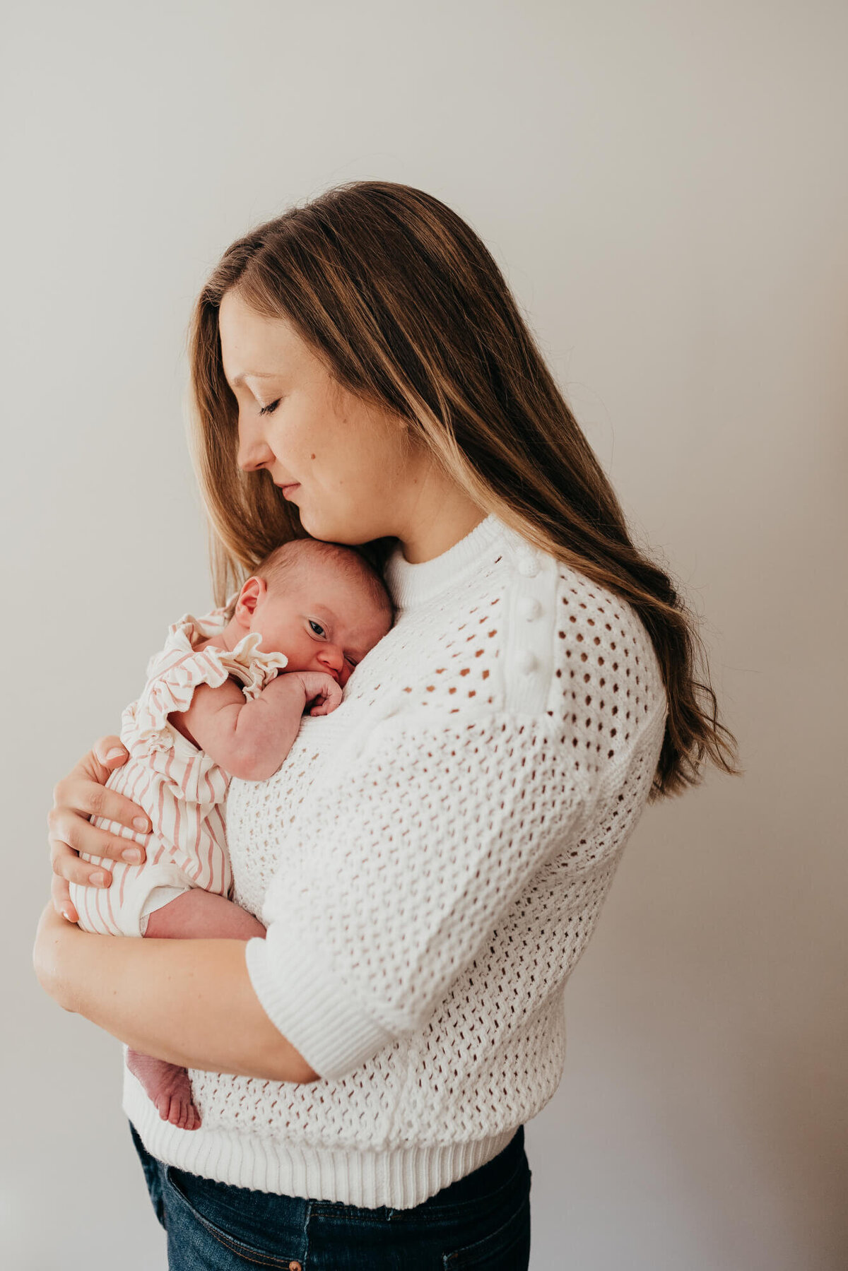 A young mother holds her newborn baby girl against her chest in front of a plain white wall