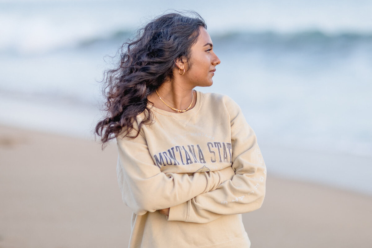 A young woman with long, wavy brown hair wearing a beige crewneck that says "Montana State," looking to her left at the ocean in Newport Beach.