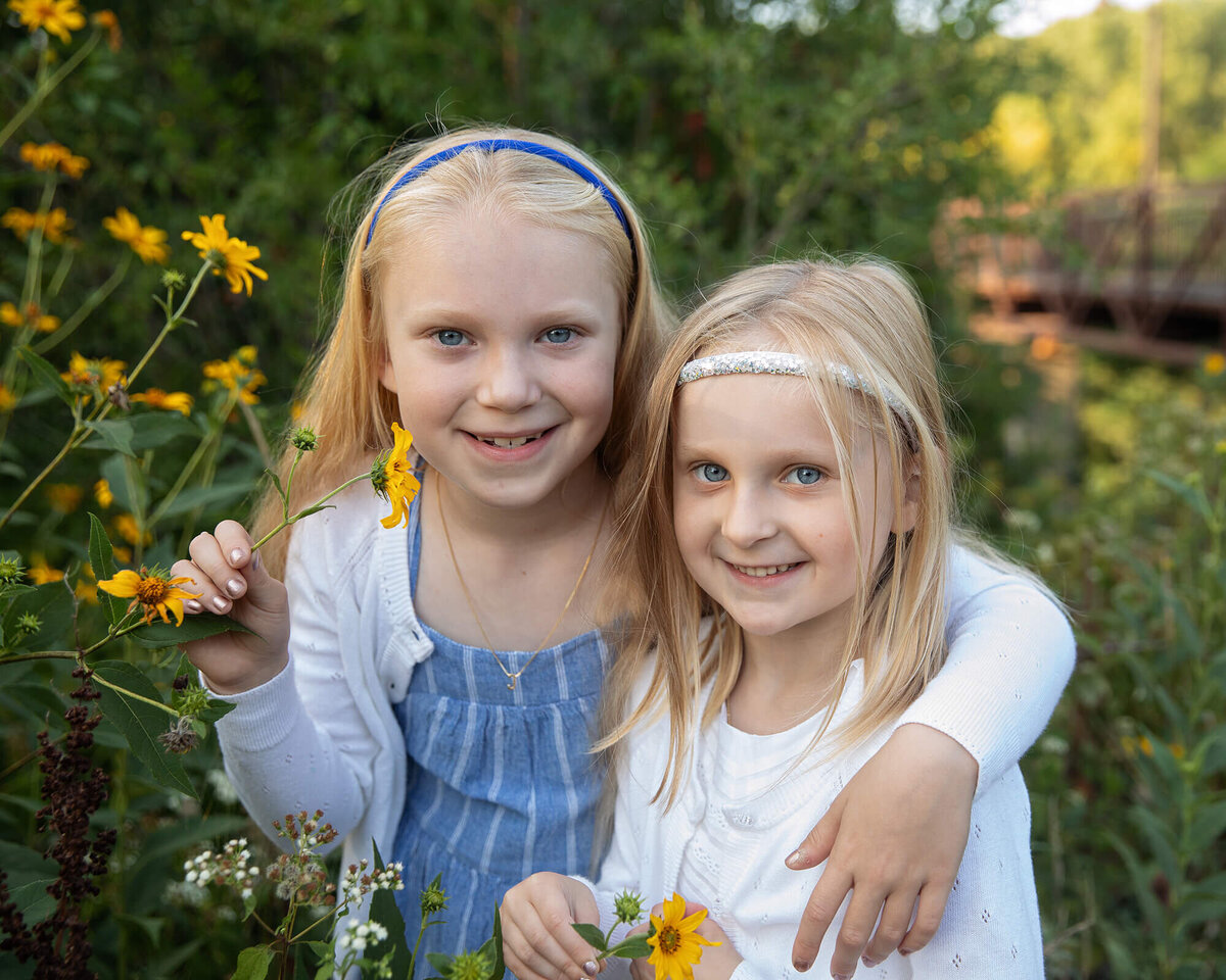 Elementary school sisters with their arm around each other in front of flowers.
