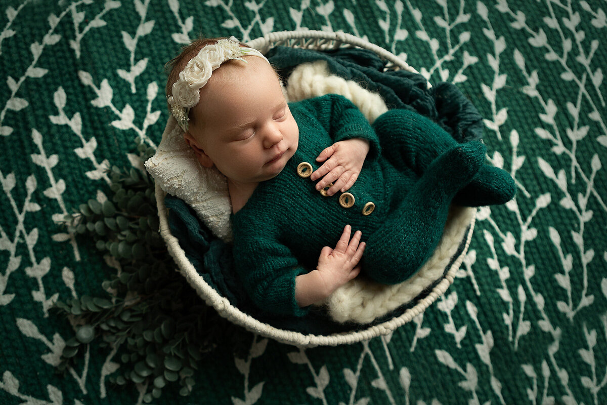 best-columbus-ohio-newborn-photographer-baby-girl-in-emerald-green-footed-sleeper-curled-in-white-basket-on-emerald-green-and-white-rug