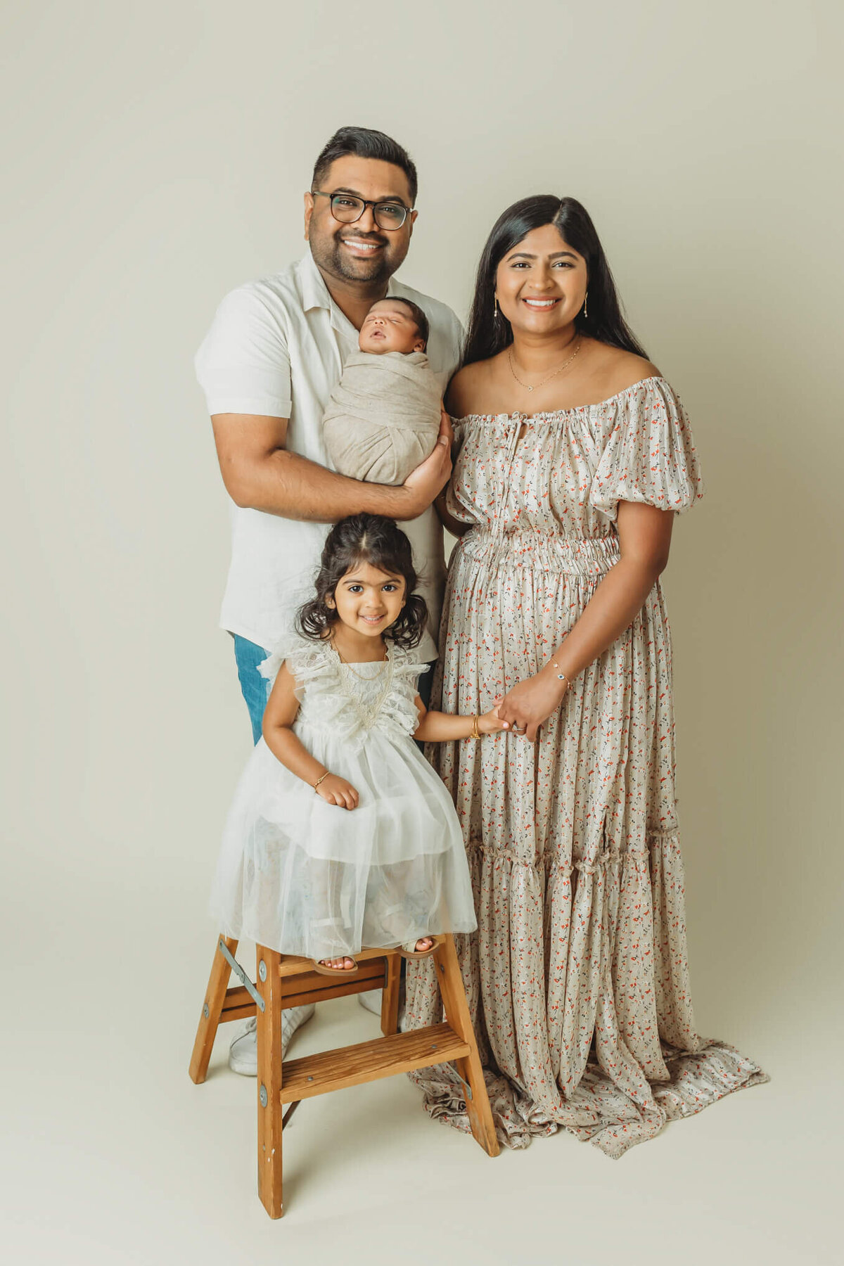 A family of four poses together for a newborn session. The father holds a wrapped newborn baby, while the mother stands beside him with a gentle smile. Their older daughter, dressed in a white dress, stands on a stool next to them, looking joyfully at the camera. The background is light and neutral, contributing to a warm and inviting atmosphere.