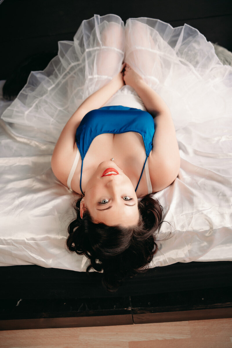 woman with dark hair  in a pinup style wearing a blue top and white crinoline skirt on a bed in the studio in Minneapolis mn