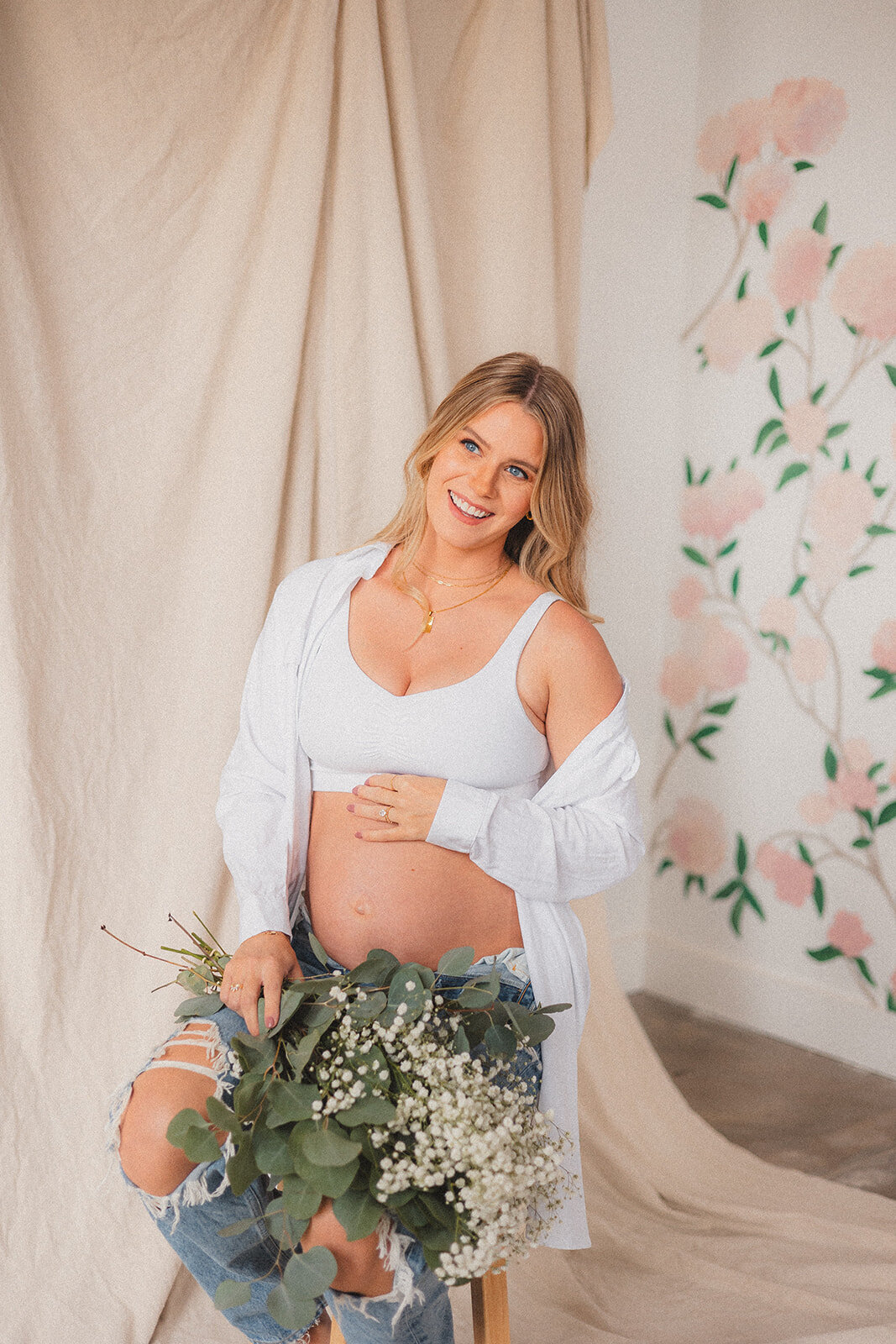 A pregnant woman poses on a stool with jeans while holding flowers