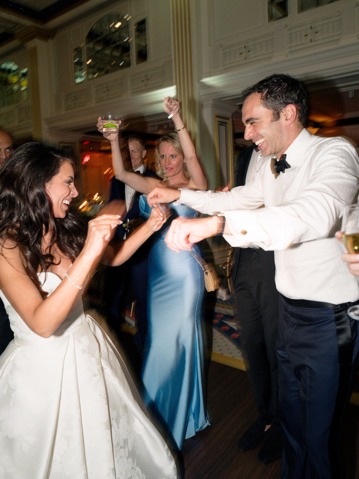 A bride and groom joyfully dancing together at their wedding reception. The bride is in a white dress, and the groom is in a white shirt with a bow tie. Guests in elegant attire surround them, raising glasses in celebration.