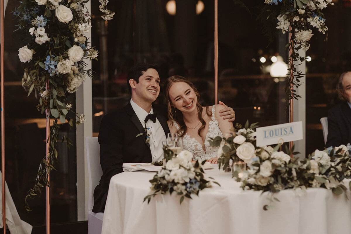 Newlyweds sitting at a reception table with their arms around each other 