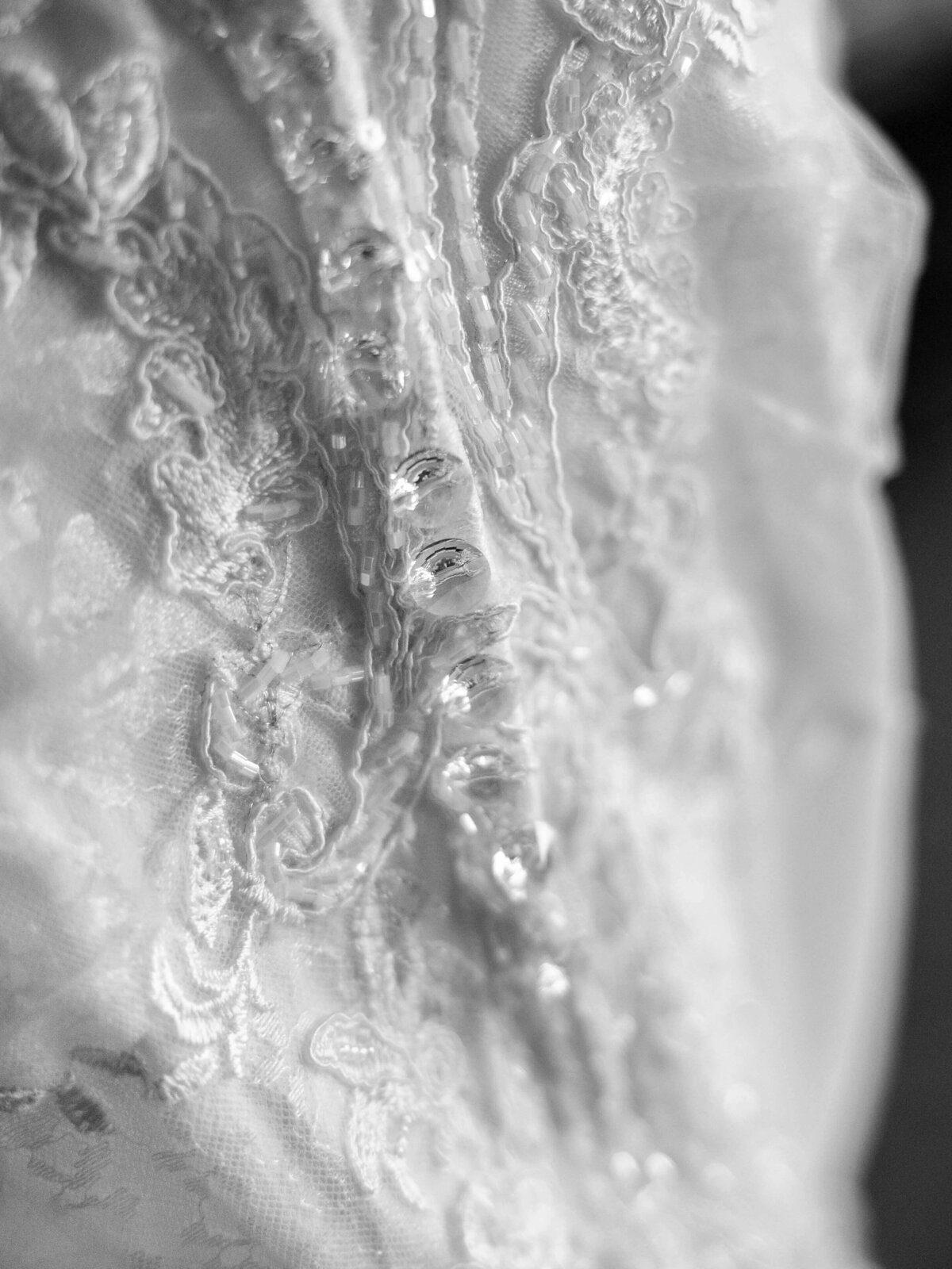 Black and white close-up of an intricately detailed lace wedding gown. The fabric features delicate floral patterns and shimmering sequins, creating a textured and elegant appearance.