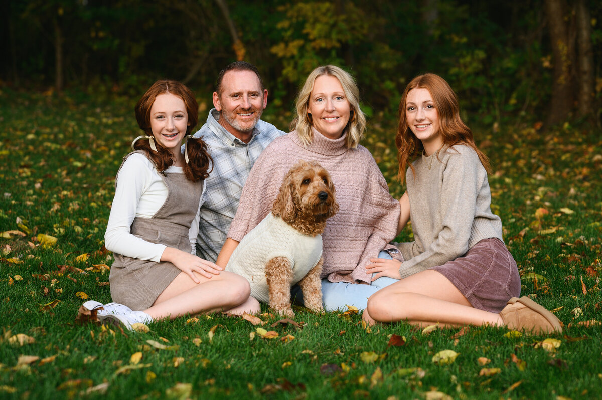 A family of four and their dog, sitting together on the grass surrounded by autumn leaves, smiling for the camera