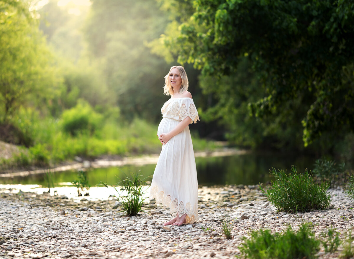 Pregnant woman in a boho white dress smiling beside a rocky creek during golden hour in Kyle, TX
