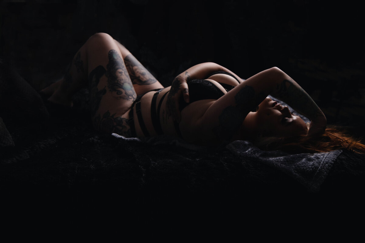 A person with tattoos, wearing black lingerie, lies on their side in a dark and moody setting, with arms raised over their head. The lighting is low, casting shadows and emphasizing their figure and inked designs.