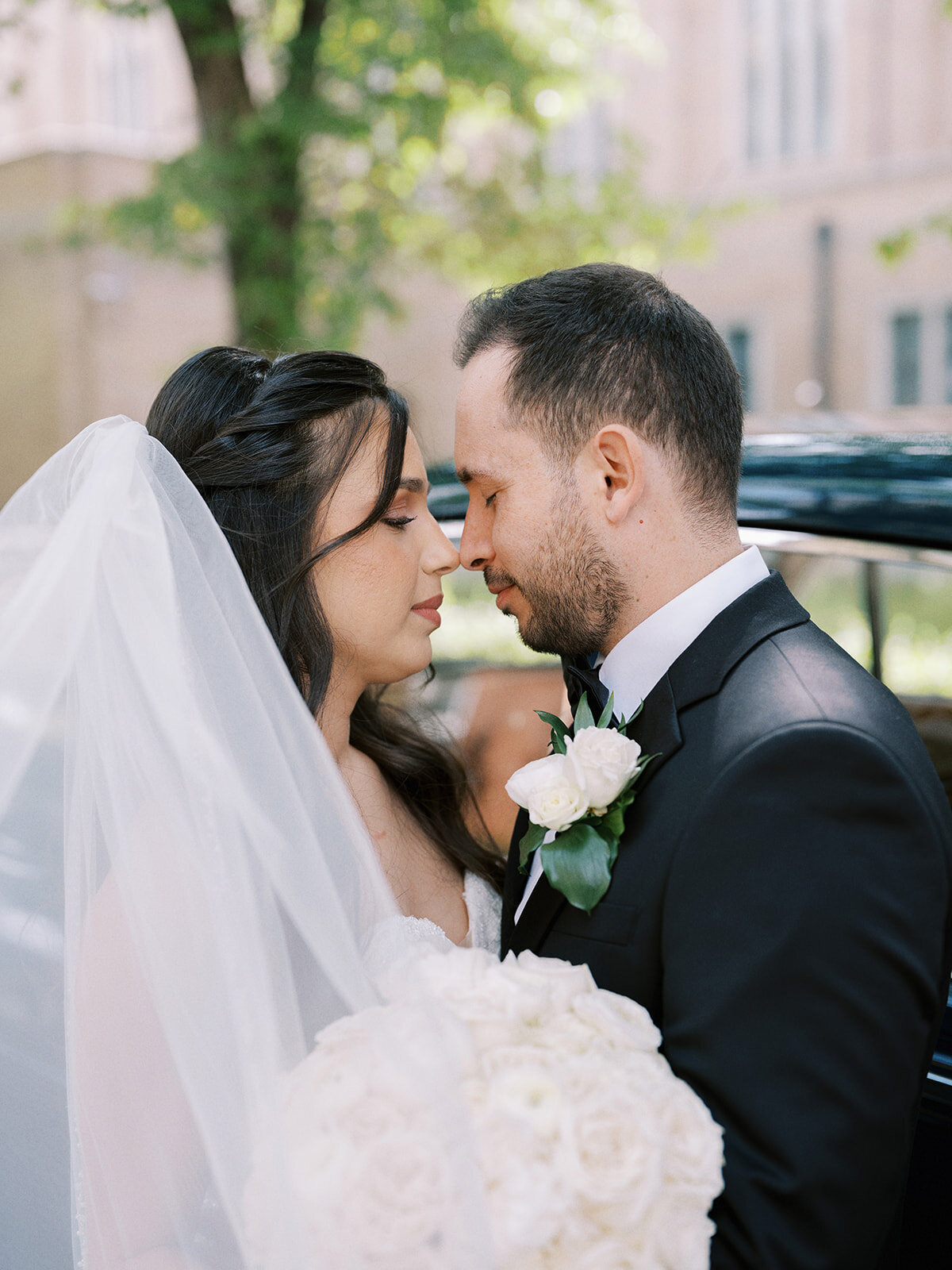 A bride and groom stand in close embrace, facing each other with eyes closed outside on a sunny day at their Fairmont Palliser Wedding in Calgary. The bride holds a bouquet of white flowers and wears a veil, while the groom is dressed impeccably in a black suit.