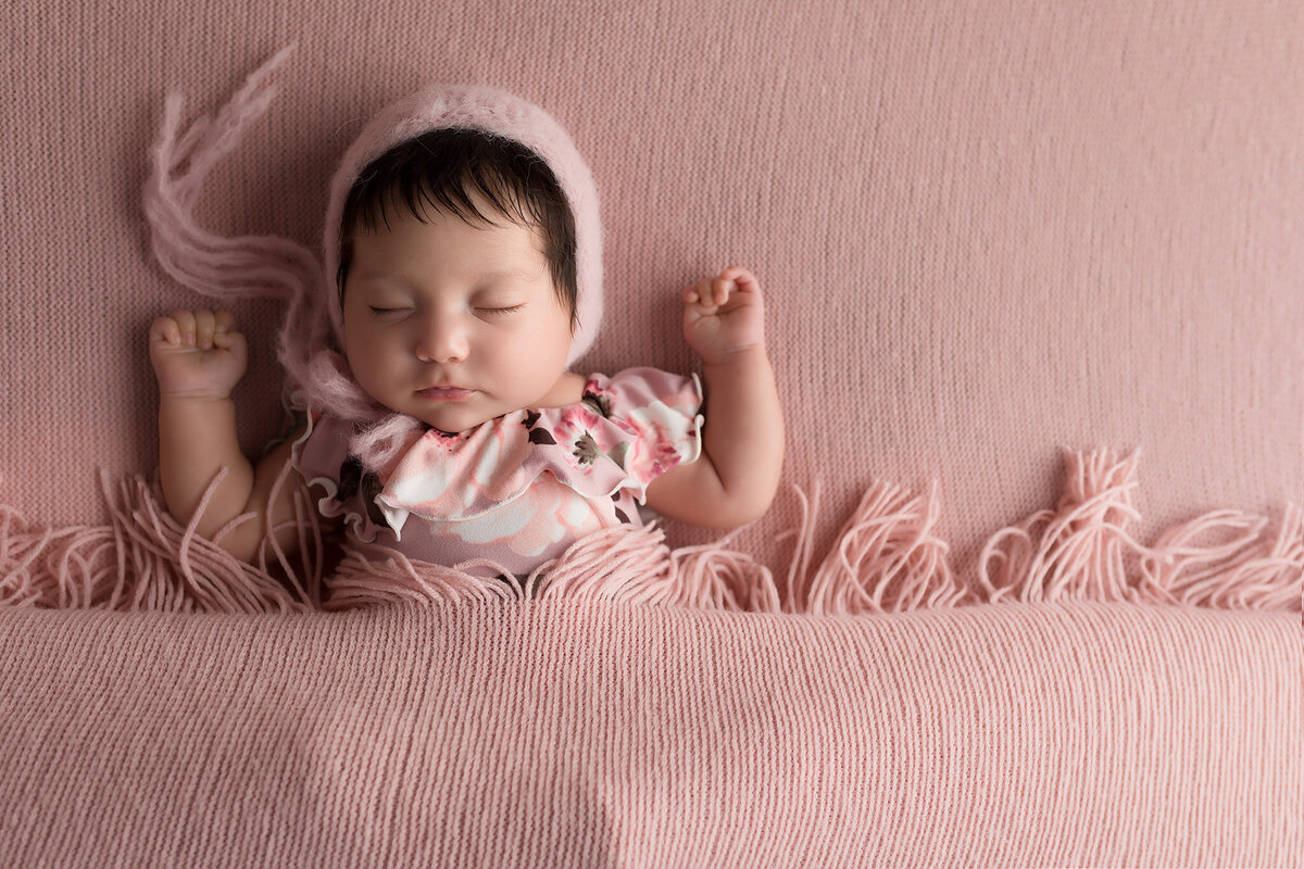 Newborn baby girl dressed in a floral pink romper with a knit bonnet, resting on a textured pink blanket with fringe, captured by Dallas newborn photographer.