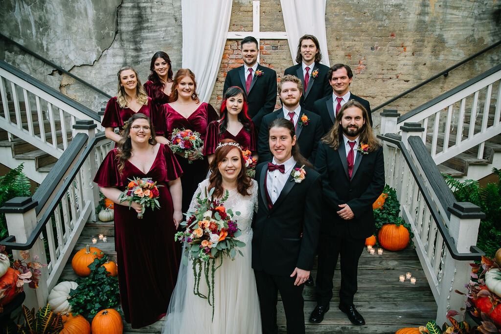 A bride and groom stand with their wedding party on outdoor stairs, surrounded by pumpkins, ferns, and candles. Bridesmaids wear burgundy dresses; groomsmen wear dark suits with red accents. A white drape hangs behind them.