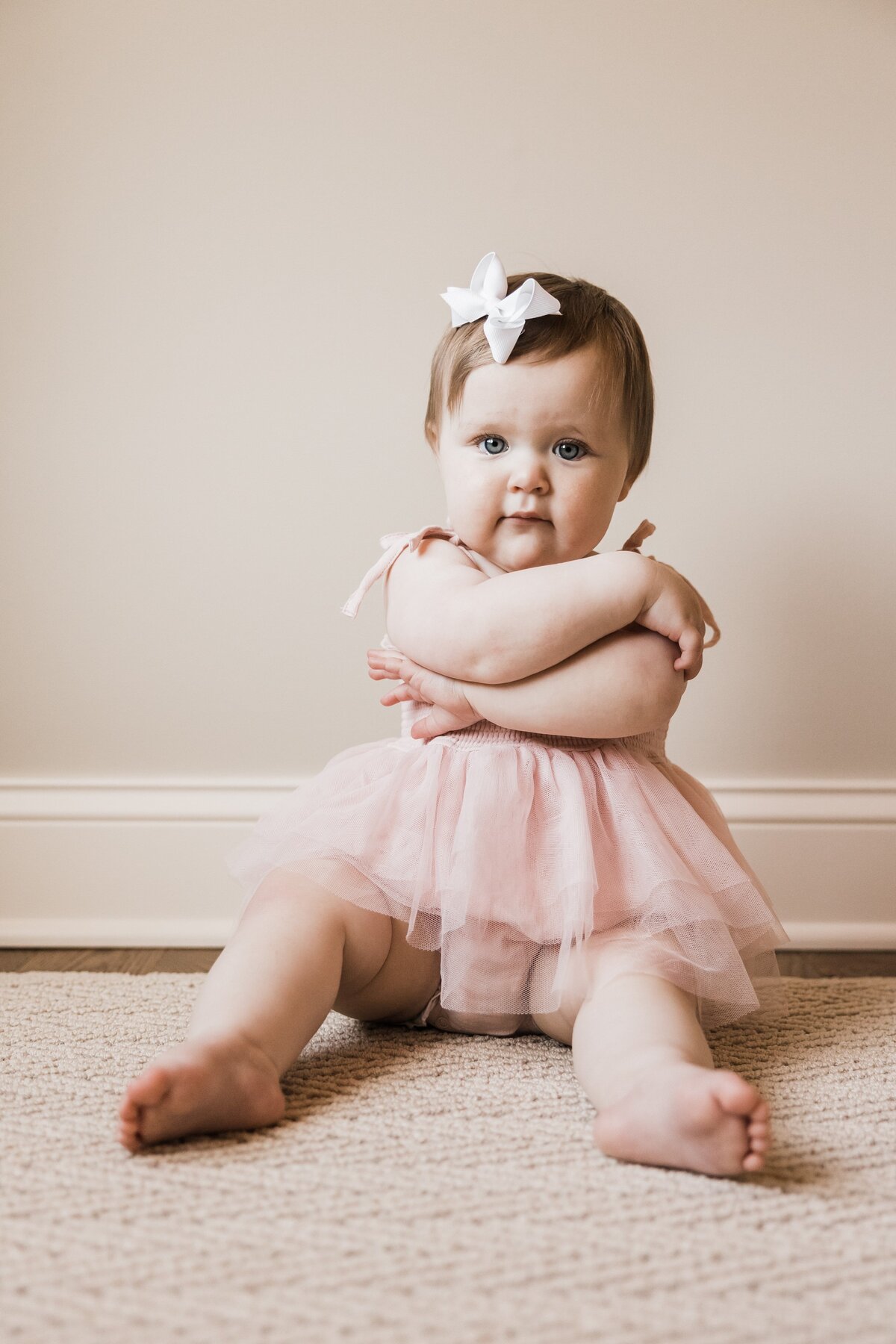 A baby girl wearing a pink tutu and a white bow headband sits on a carpeted floor, ready for her session with the family photographer in Pittsburgh, PA.