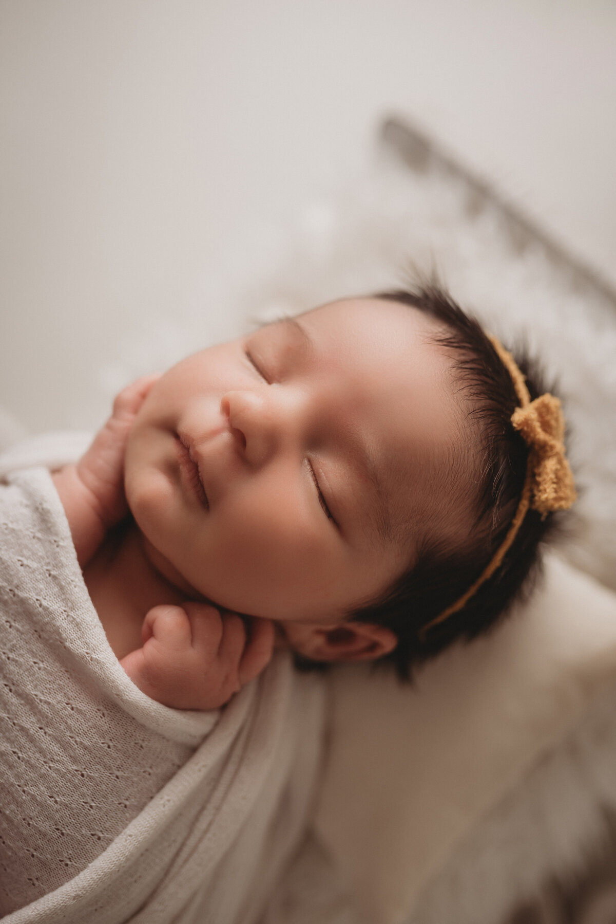 Smyrna, GA newborn photographer photographed sleeping newborn girl with hands on cheeks, dressed in off white swaddle and yellow headband with bow