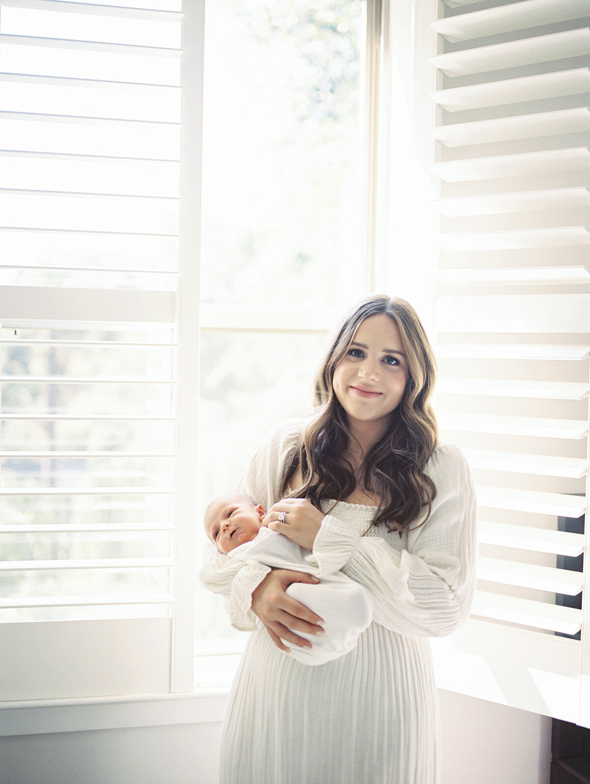 Mother with brown hair holds newborn baby while standing in front of the open window in her home, smiling at the camera in her Bethesda, Maryland home.