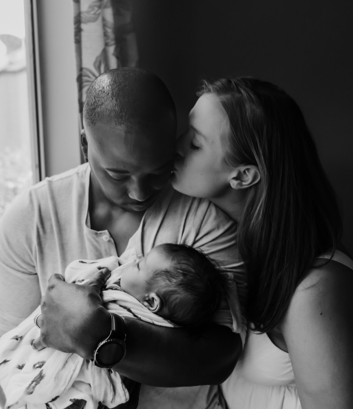 Newborn Photographer, Mom and dad stand by the window and snuggle close together and kiss.
