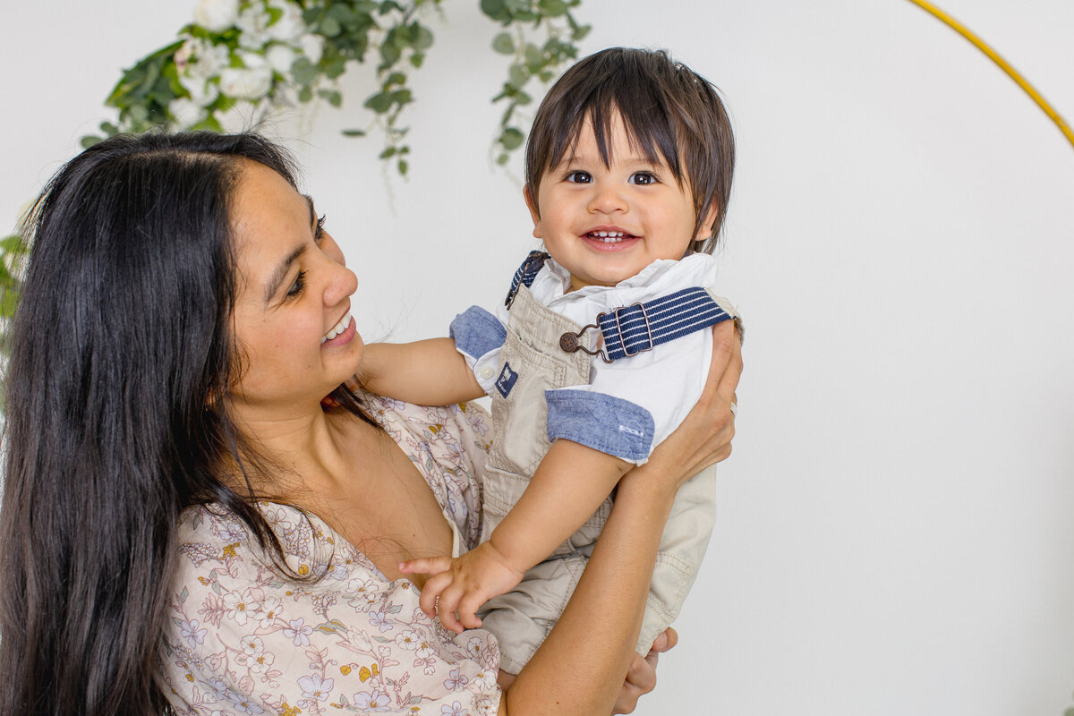 A mom with long, black hair smiling at her young son as he smiles and looks at the camera in her arms. Captured in a studio in Tustin.