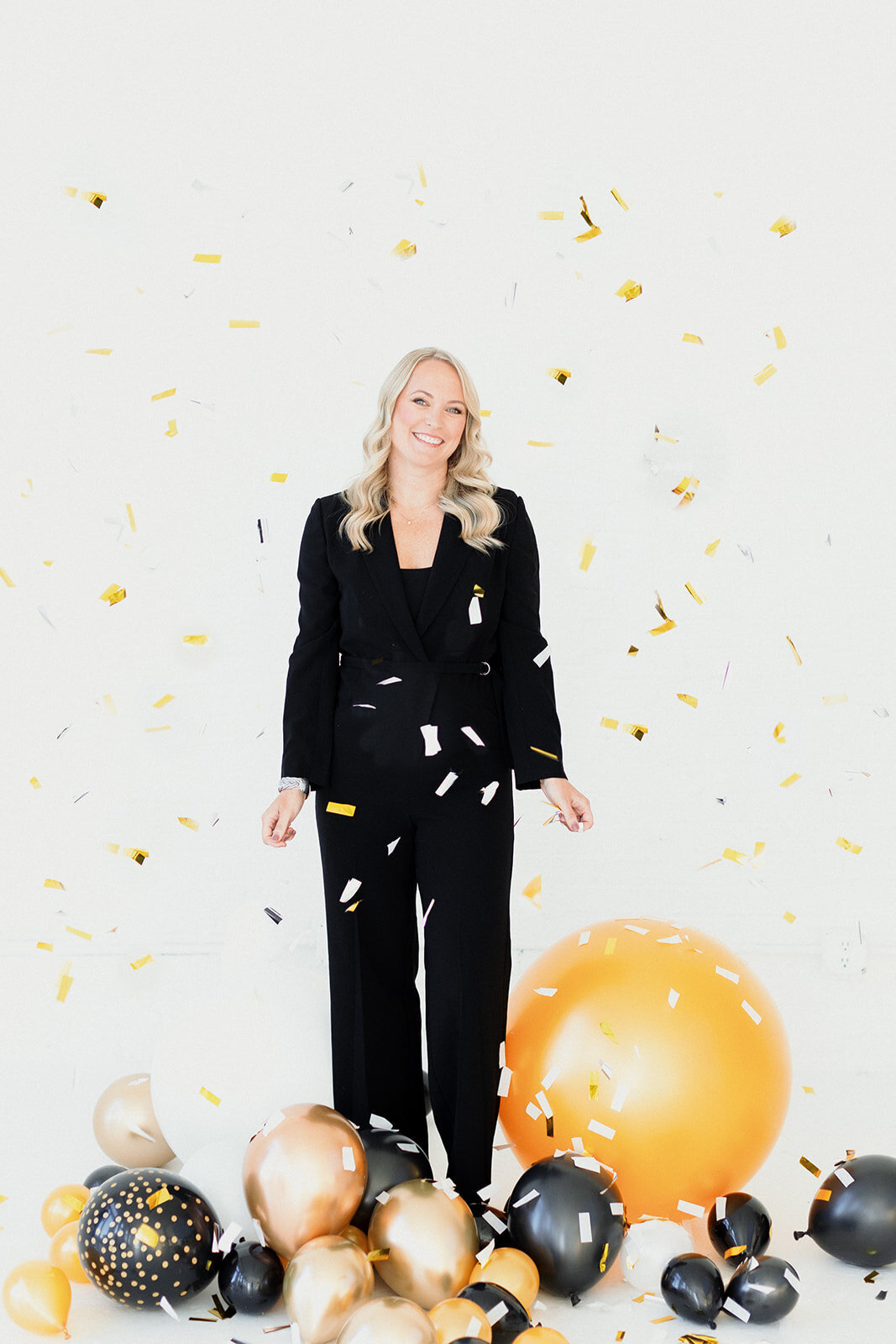 blonde woman stands in front of a white backdrop with black and gold balloons at her feet as gold confetti falls from the sky. She is smiling and wearing a black suit