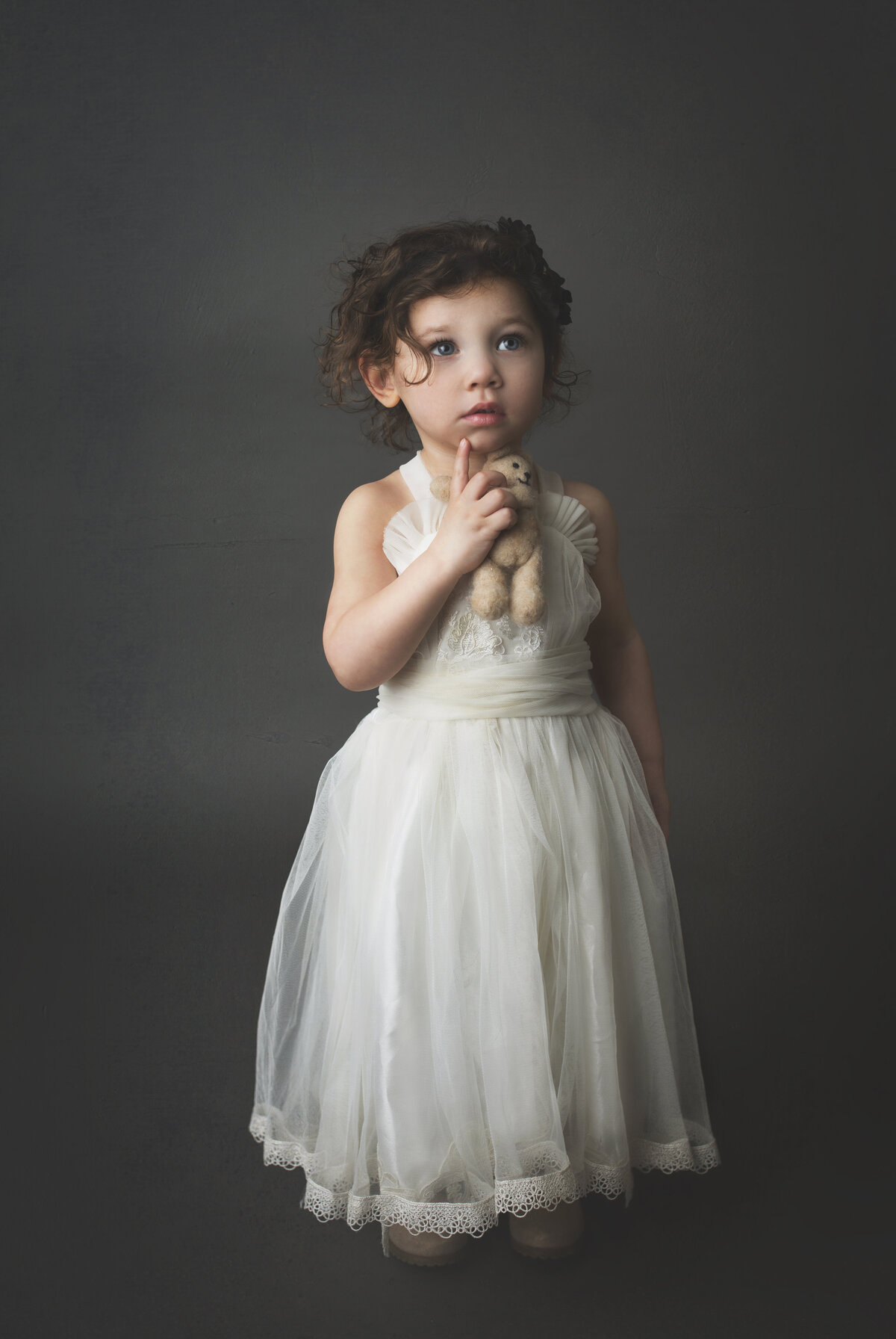 Toddler girl standing in white dress with grey background, holding a teddy bear