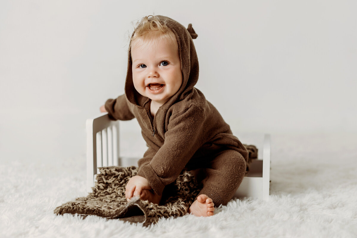 Happy six month old baby boy sits on newborn sized bed wearing a brown knit bear outfit during his Denver milestone session