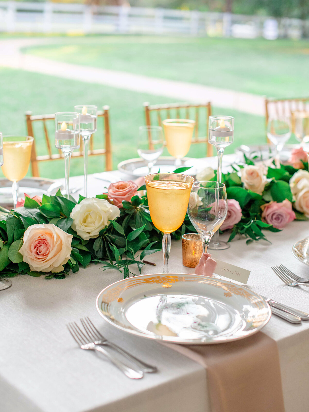 A beautifully set table with elegant dinnerware, surrounded by a lush garland of pink and white roses. Yellow-tinted glasses and several candles in glass holders adorn the table, with a garden visible in the background.