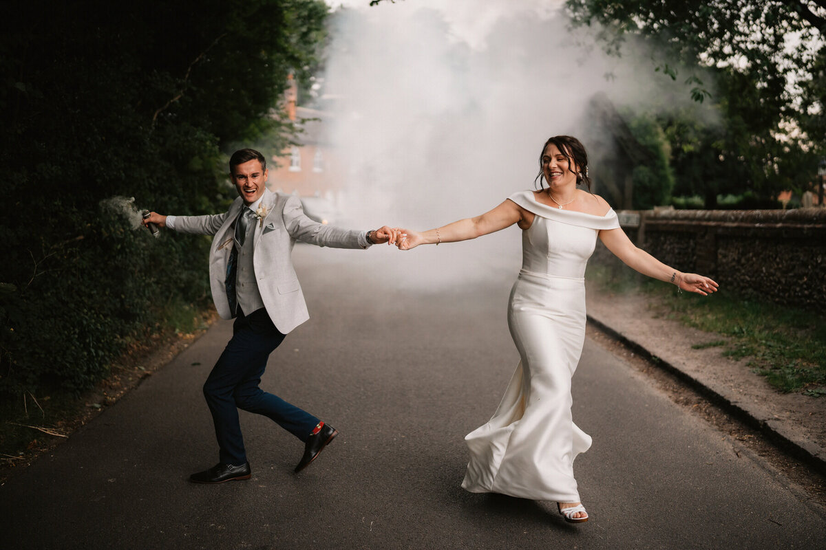 Bride and groom joyfully running hand in hand down the road outside The Cock Inn, Sarratt, surrounded by smoke and laughter during their luxury countryside wedding.