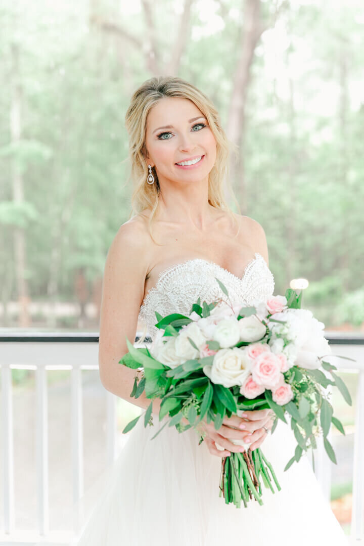 A happy bride stands on a porch smiling with her pink and white bouquet