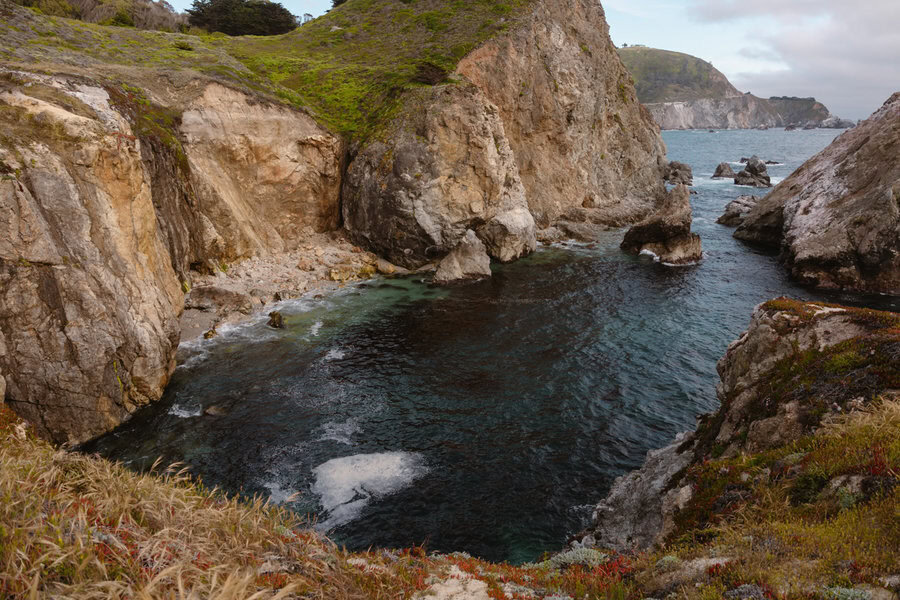 A rocky coastal inlet with clear blue water, surrounded by steep cliffs and lush green vegetation.