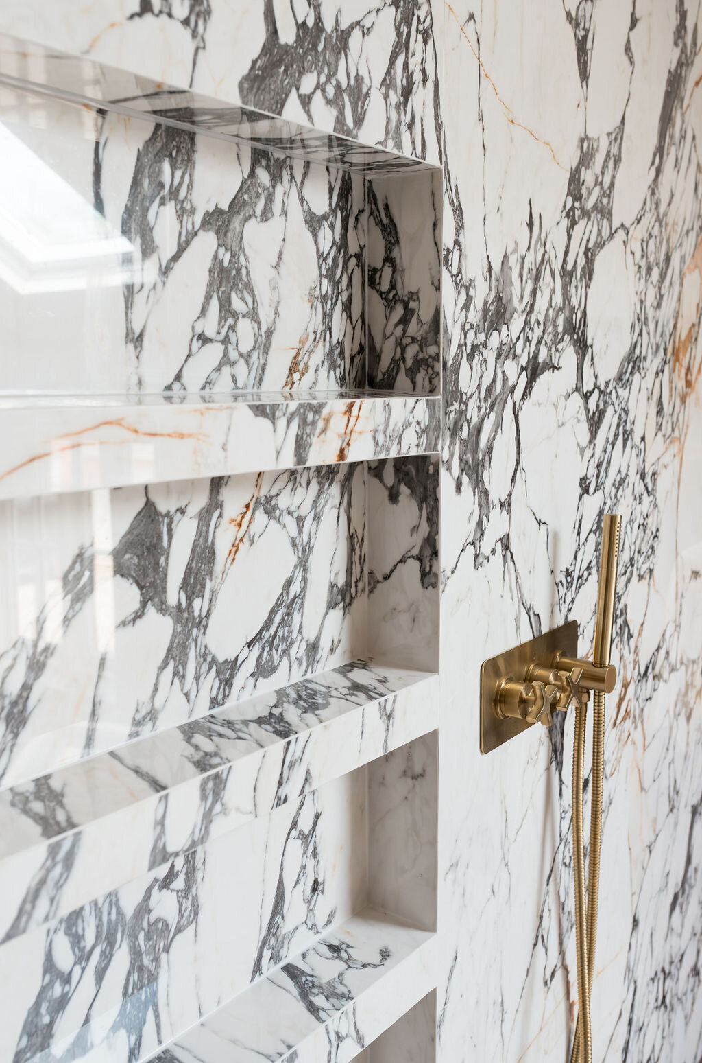 Modern gold shower fixtures mounted on a white marble wall with black veining. To their left, multiple shelves are built into the marble wall.