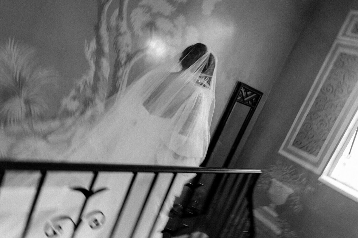 A black-and-white bridal portrait at Millstone Adams Pond captures a bride ascending a staircase, her veil cascading behind her in soft, flowing layers. The motion blur adds an ethereal, dreamlike quality, emphasizing the movement and emotion of the moment. The intricate wall art and architectural details frame the composition beautifully, creating a timeless and artistic wedding image.