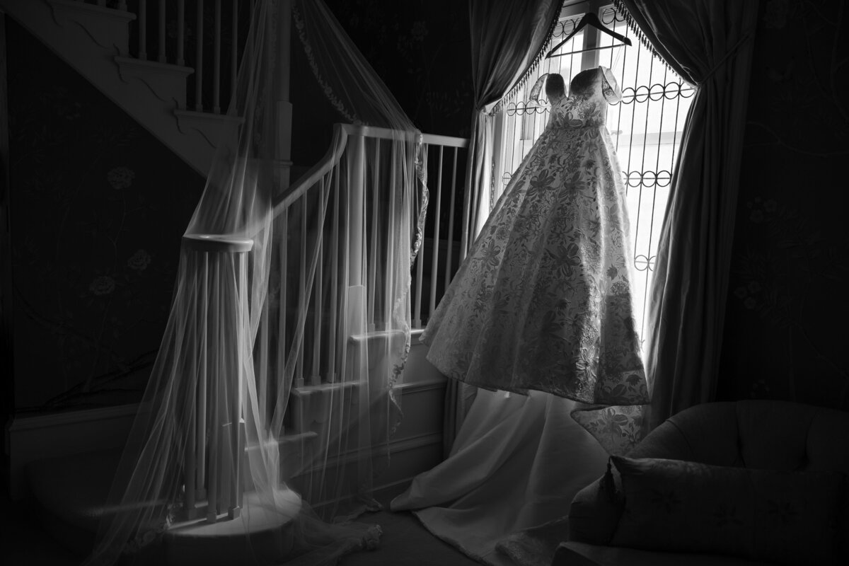 A wedding gown with floral patterns hangs in a window framed by curtains, while a long embroidered veil is draped over the banister of a curved staircase, both softly lit by natural window light in a quiet, elegant room.