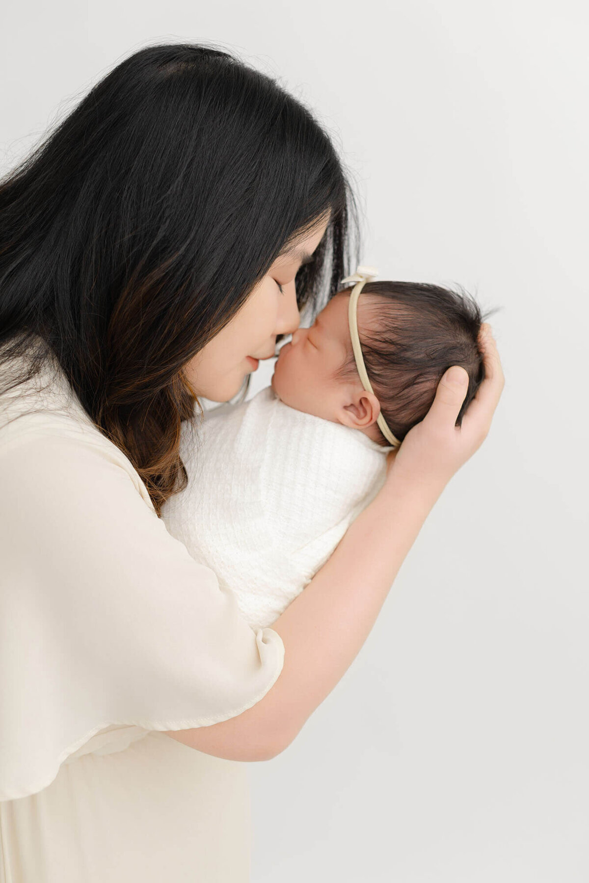 Mom in cream dress holding baby up to her nose so that their noses are touching. Baby is sleeping and swaddled in white.