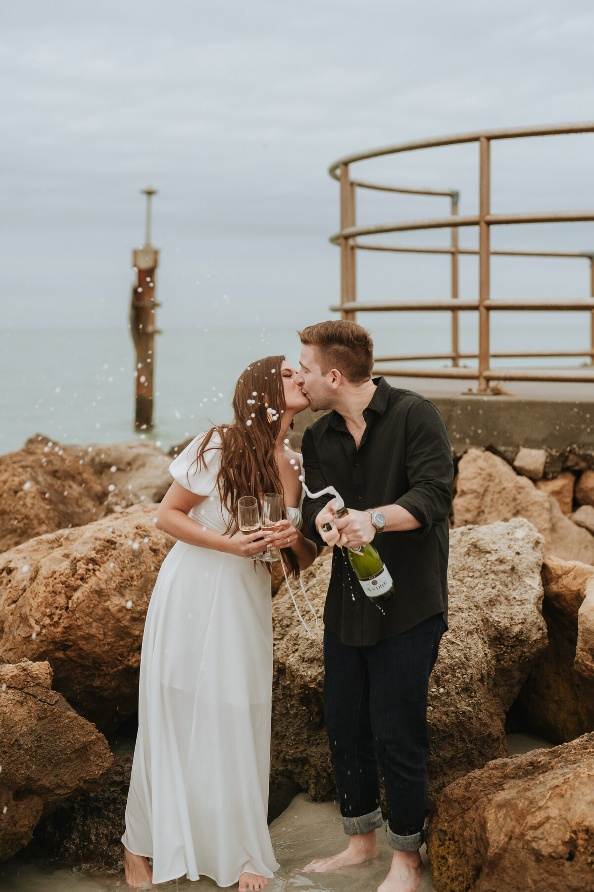 couple popping champagne on the beach while kissing during engagement session