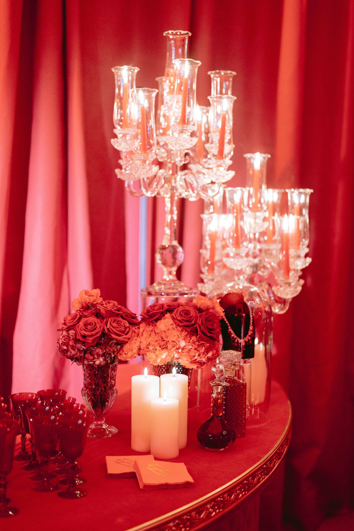 The stunning photo was taken at the prestigious Shangri La Hotel & Ritz Paris Hotel in Paris, France, by the talented photographer Matteo Coltro, with videography by Zen Film Works. The scene captures a lavish table adorned with elegant glass candelabras, each holding flickering lit candles that cast a warm and inviting glow. The table is beautifully decorated with clusters of vibrant red roses, exquisite decorative glassware, and a couple of large white candles adding to the elegant atmosphere. The backdrop of rich red drapes further enhances the romantic ambiance, making it a truly enchanting and exquisite setting.
