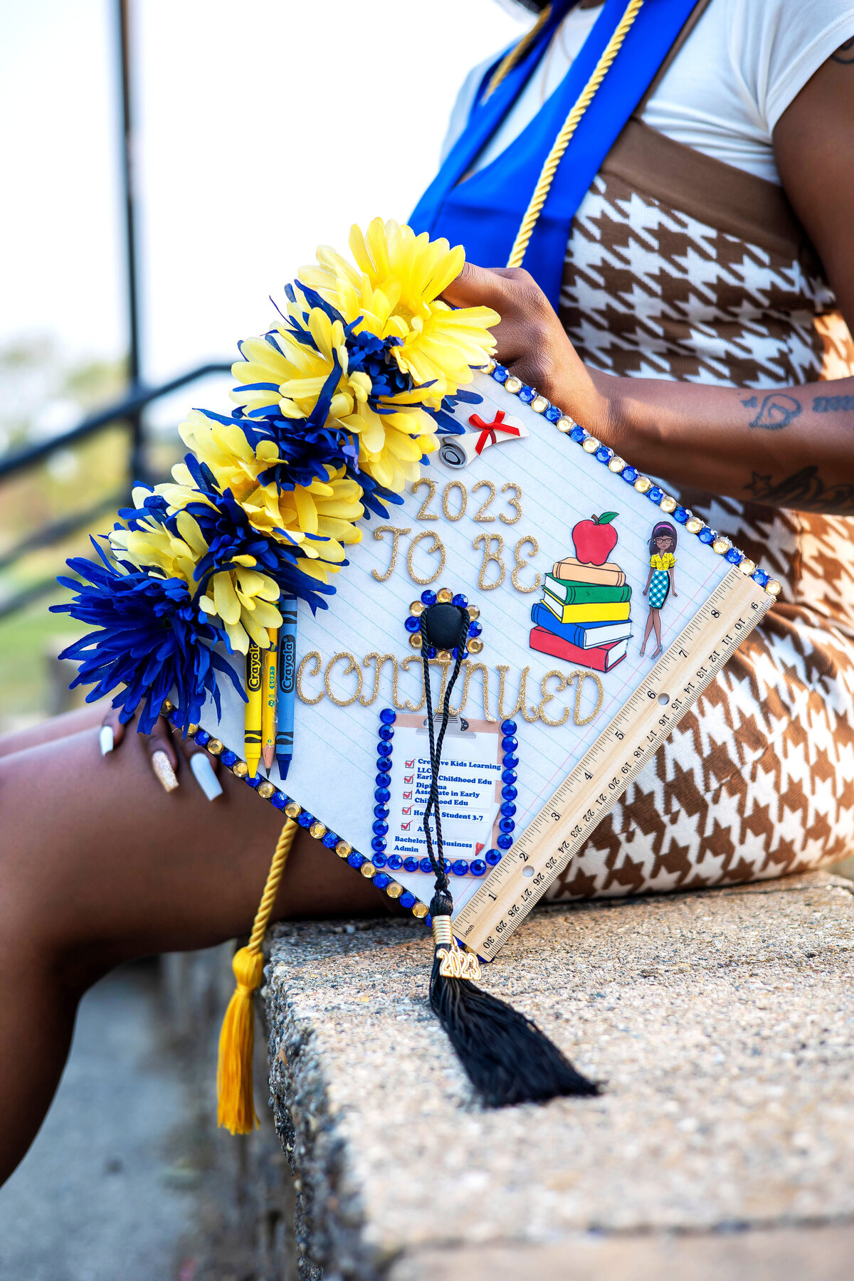College graduate wearing brown and white dress sitting on ledge holding decorated graduation cap