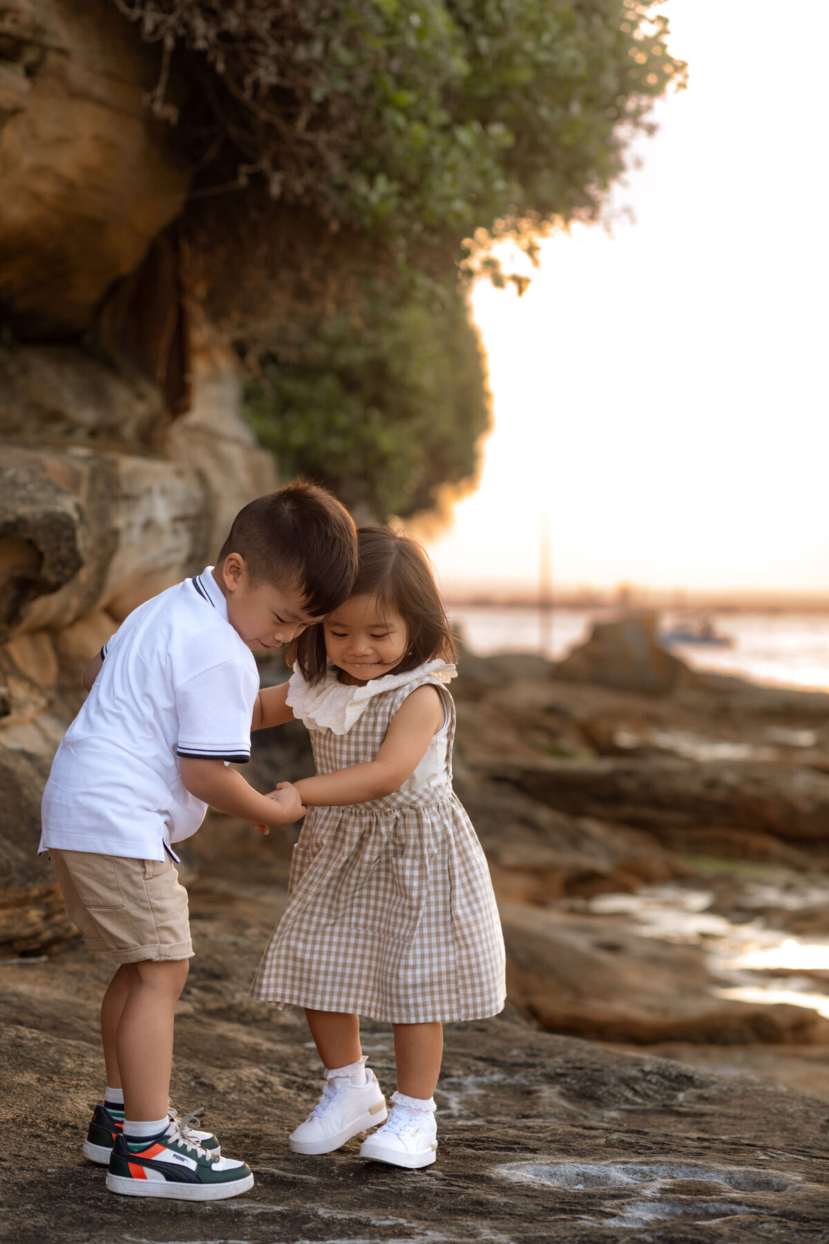 Young brother and sister sharing a sweet moment by the water at sunset, holding hands and smiling during a relaxed Sydney family photo session.
